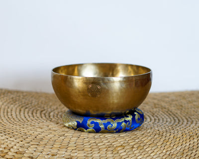 Angled top view of a hand-hammered golden bronze singing bowl displaying an engraved tiger interior design, shown next to a suede-wrapped wooden striker and blue cushion.