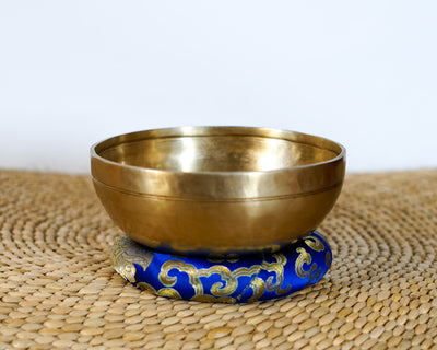 Side profile view of a small warm golden hand-hammered bronze singing bowl with double rim lines, displayed on a blue brocade cushion.