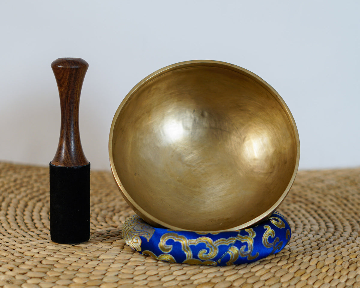 Side profile view of a small warm golden hand-hammered bronze singing bowl with double rim lines, displayed on a blue brocade cushion.