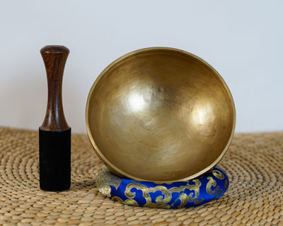 Side profile view of a small warm golden hand-hammered bronze singing bowl with double rim lines, displayed on a blue brocade cushion.