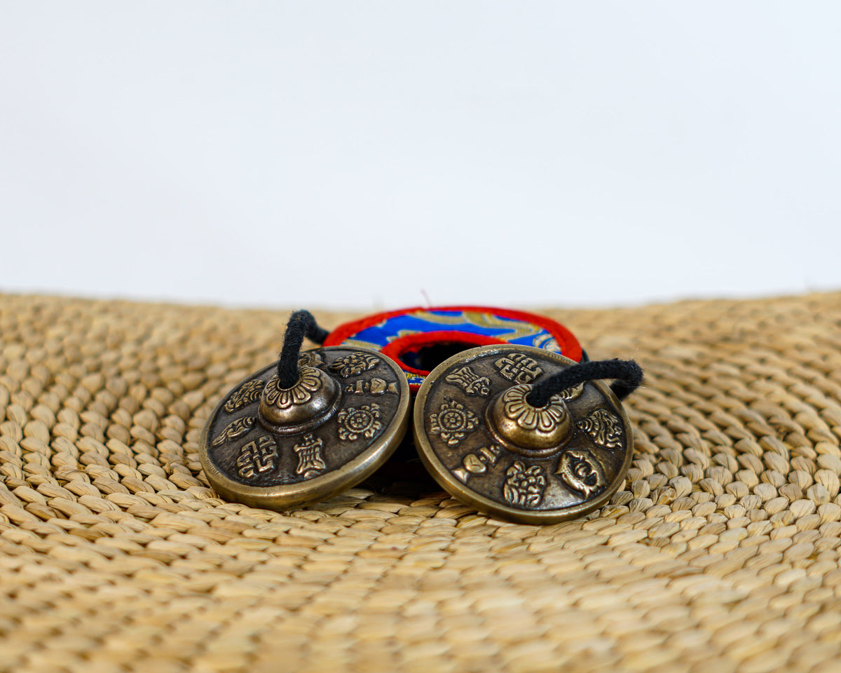 Tibetan tingsha cymbals engraved with eight auspicious Buddhist symbols and a blue brocade carrying case.