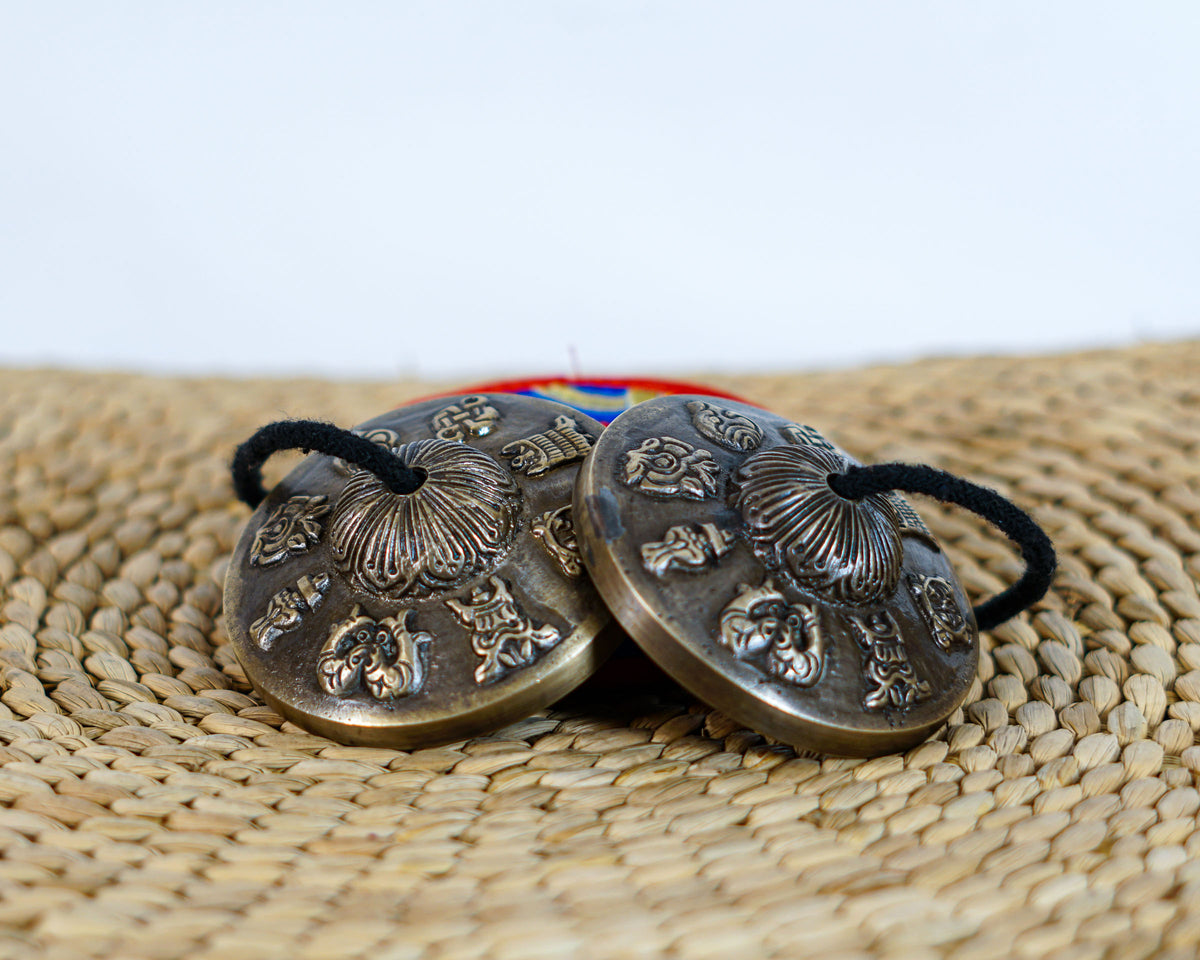 Tibetan Buddhist tingsha cymbals with detailed embossed symbols and an antique metallic finish, photographed with a colorful brocade case.