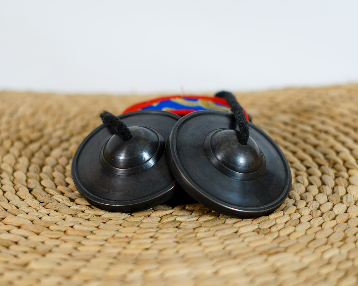 Black Tibetan tingsha cymbals with a dark metallic finish and black connecting cord displayed with a colorful brocade case.