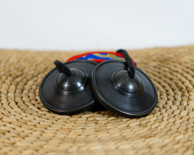 Black Tibetan tingsha cymbals with a dark metallic finish and black connecting cord displayed with a colorful brocade case.