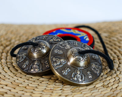 Tibetan Buddha tingsha cymbals featuring detailed Buddha engravings, a black connecting cord, and a colorful brocade carrying case.