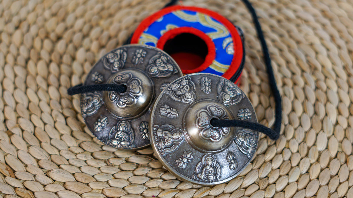Close up of Tibetan Buddha tingsha cymbals showing embossed Buddha figures and traditional decorative motifs on a dark metallic finish.
