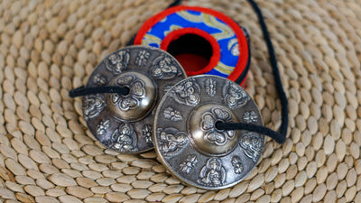 Close up of Tibetan Buddha tingsha cymbals showing embossed Buddha figures and traditional decorative motifs on a dark metallic finish.