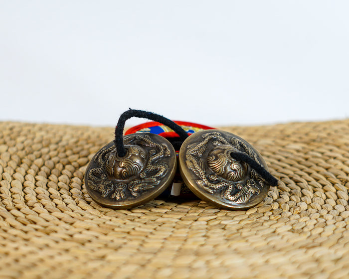 Tibetan Dragon Tingsha Cymbals featuring intricate dragon engravings, a black connecting cord, and a colorful brocade carrying case.