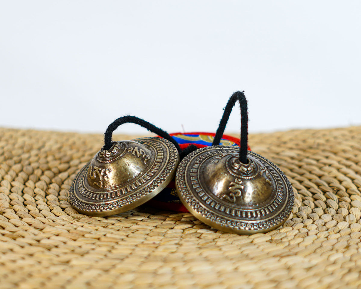 Tibetan Lotus Tingsha Cymbals with intricate concentric engravings, a black connecting cord, and a colorful brocade carrying case.