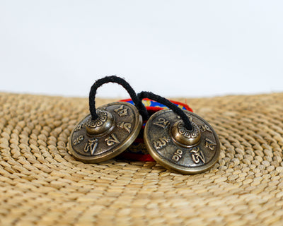 Tibetan Mantra Tingsha Cymbals with raised mantra engravings, antique metallic finish, and black connecting cord.
