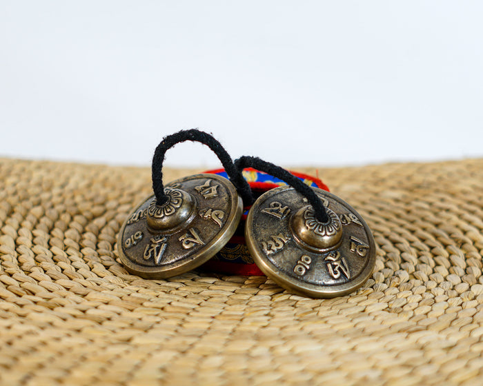 Tibetan Mantra Tingsha Cymbals with raised mantra engravings, antique metallic finish, and black connecting cord.
