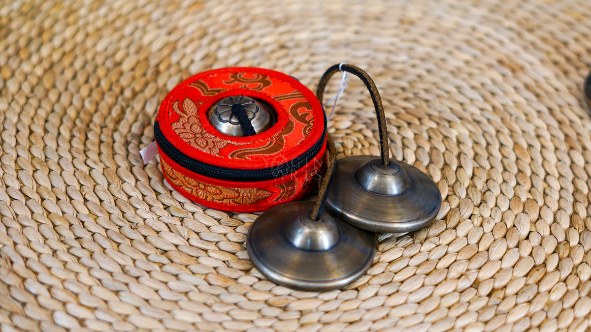 Traditional Tibetan Tingsha cymbals handcrafted from metal with a dark antique finish and connected by a cord, photographed on a natural woven surface.