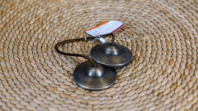Traditional Tibetan Tingsha cymbals handcrafted from metal with a dark antique finish and connected by a cord, photographed on a natural woven surface.