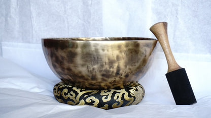 Hand-hammered brass singing bowl placed on a decorative brocade cushion with a wooden striker beside it, displayed against a white background.