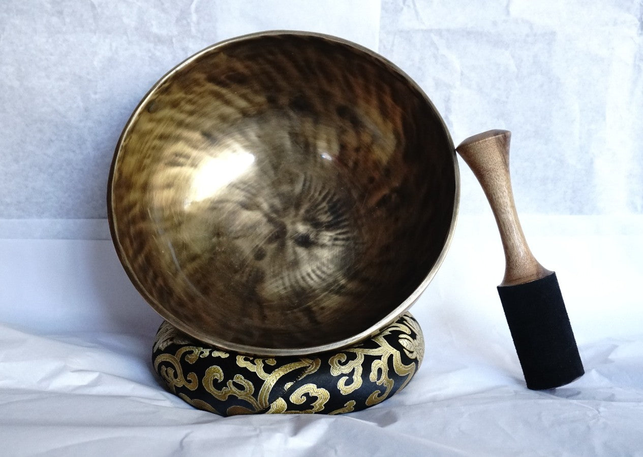 Hand-hammered brass singing bowl placed on a decorative brocade cushion with a wooden striker beside it, displayed against a white background.