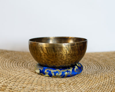 Side profile view of a small golden bronze Tiger style hand-hammered singing bowl with mottled surface patina, resting on a blue brocade cushion.