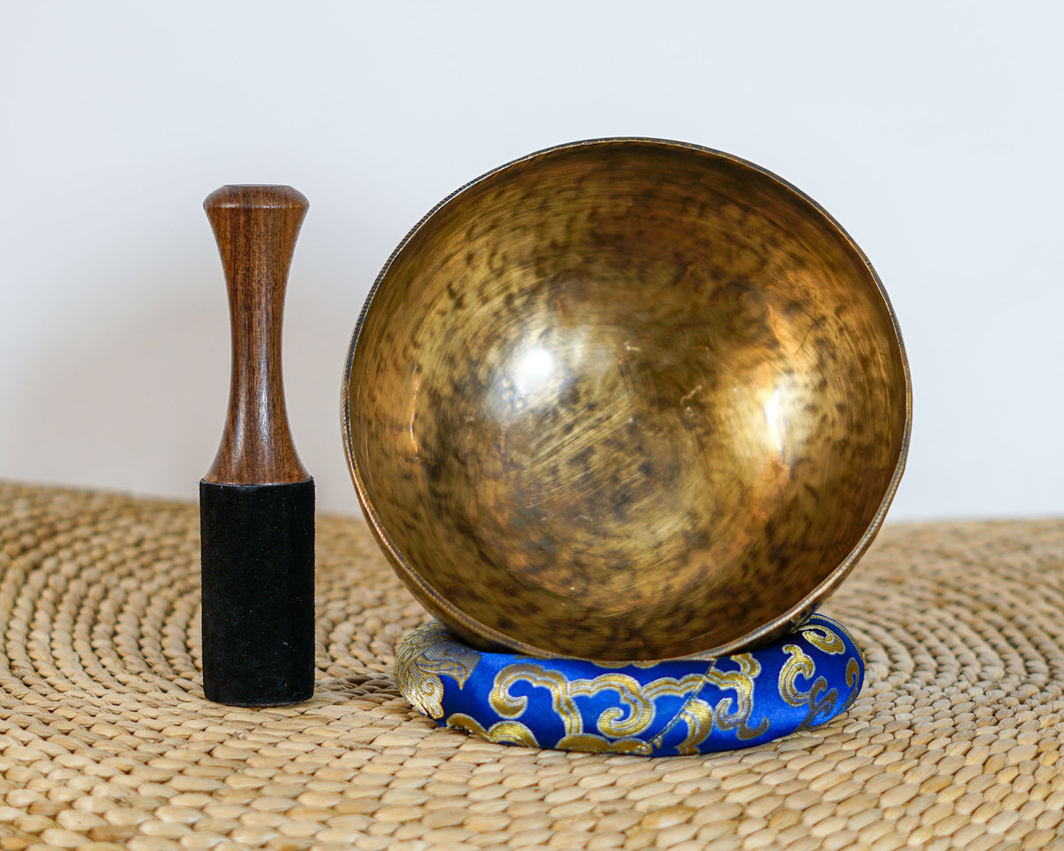 Side profile view of a small golden bronze Tiger style hand-hammered singing bowl with mottled surface patina, resting on a blue brocade cushion.