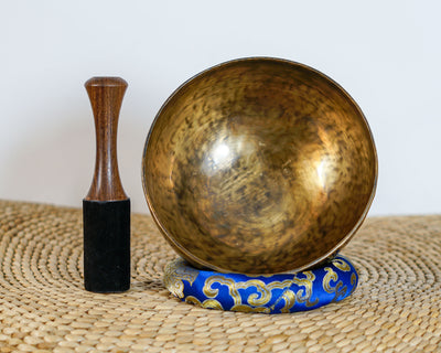 Side profile view of a small golden bronze Tiger style hand-hammered singing bowl with mottled surface patina, resting on a blue brocade cushion.