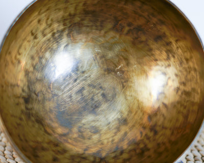 Side profile view of a small golden bronze Tiger style hand-hammered singing bowl with mottled surface patina, resting on a blue brocade cushion.