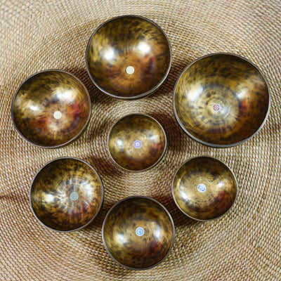 Seven handcrafted chakra singing bowls arranged together, showing different sizes, warm antique metallic finishes and individual chakra symbols.