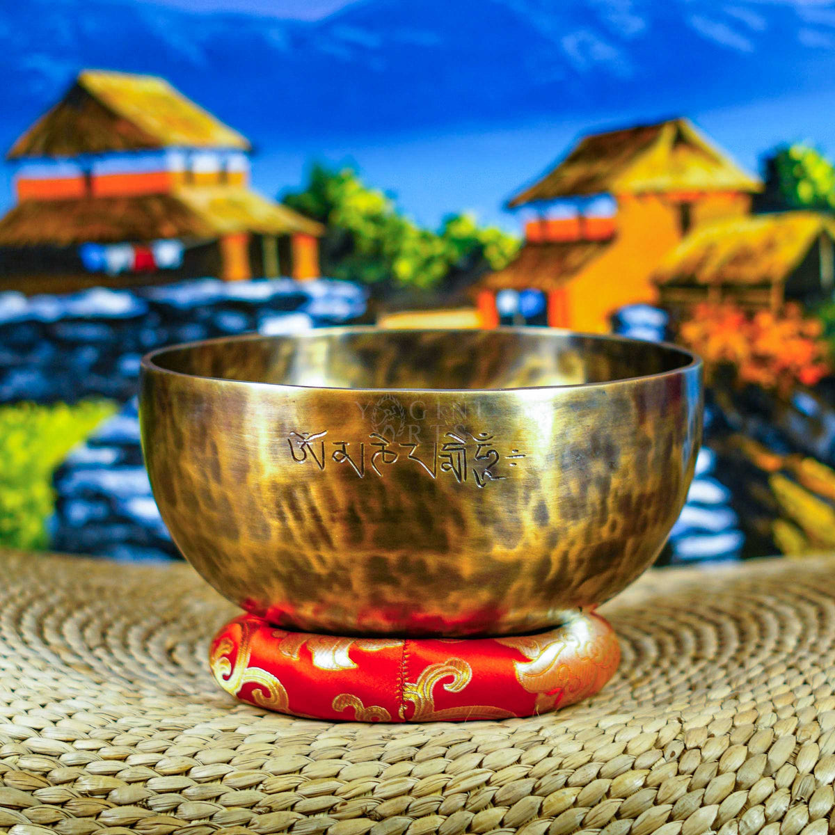 Handcrafted seven chakra singing bowl set with a brass-toned bowl, Om symbol, red cushion and wooden-handled striker displayed against a Himalayan backdrop.