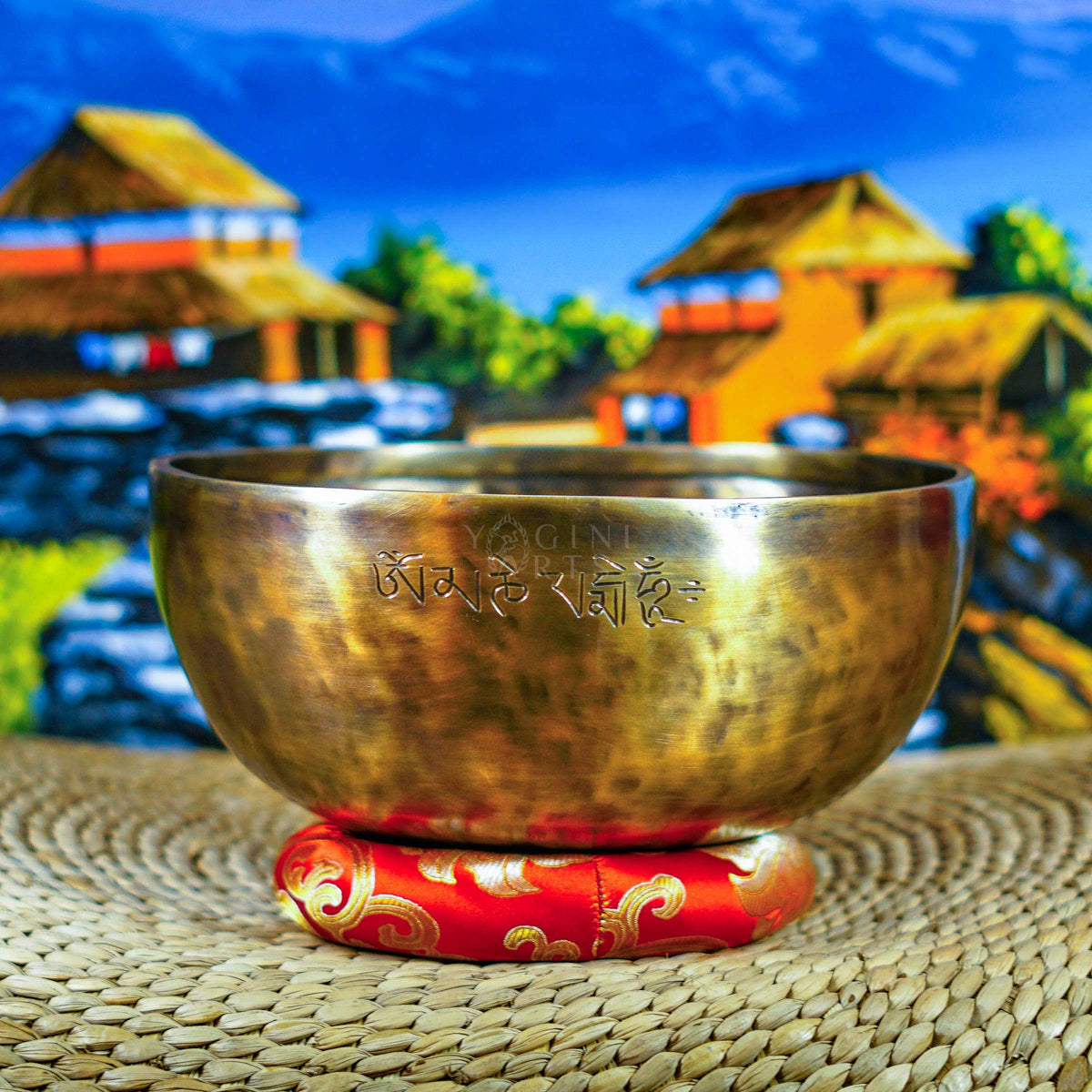 Handcrafted seven chakra singing bowl set with a brass-toned bowl, Om symbol, red cushion and wooden-handled striker displayed against a Himalayan backdrop.