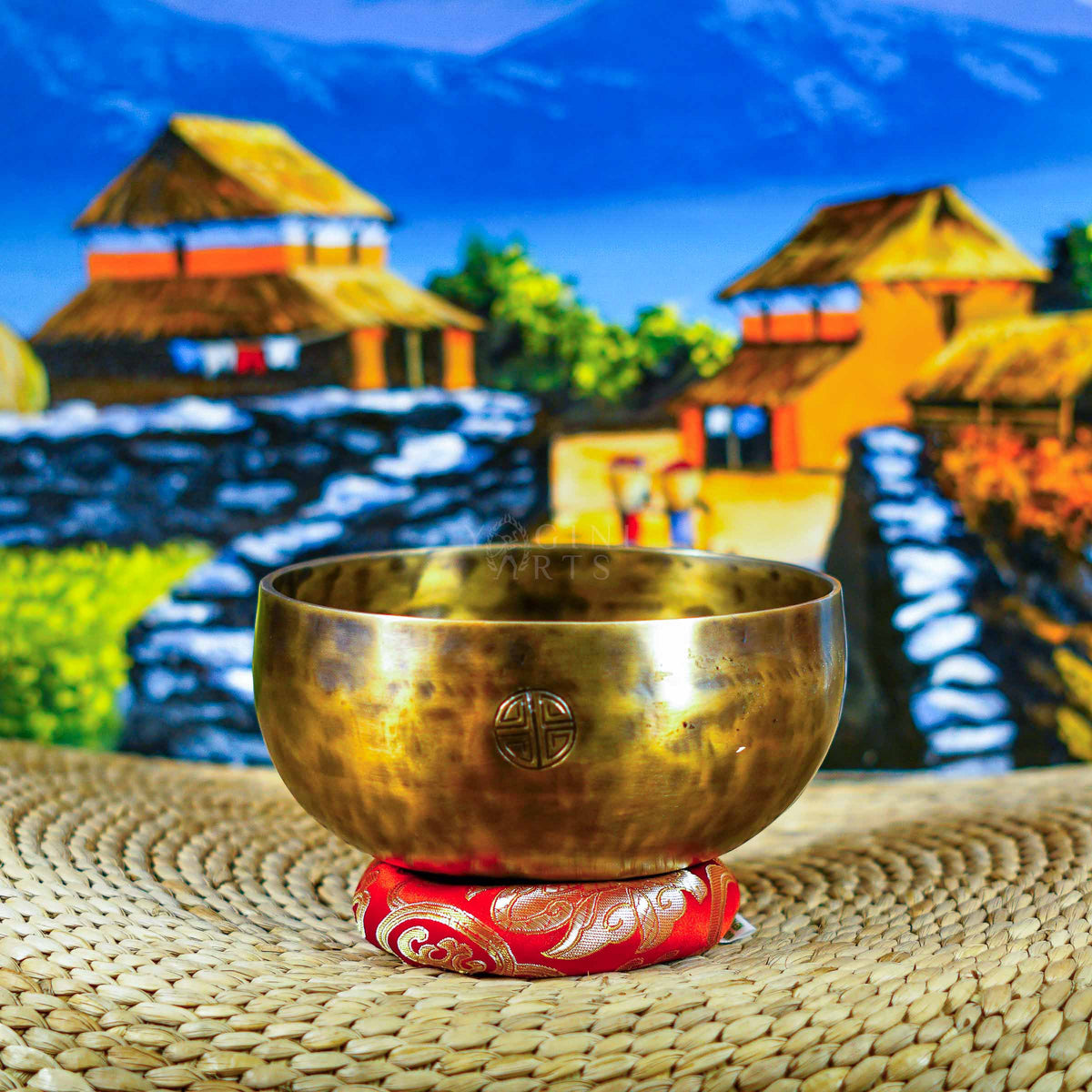 Handcrafted seven chakra singing bowl set with a brass-toned bowl, Om symbol, red cushion and wooden-handled striker displayed against a Himalayan backdrop.