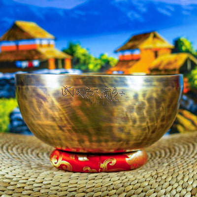 Side view of a handcrafted Himalayan singing bowl with warm bronze-gold finish, engraved Tibetan-style inscription and red decorative cushion.