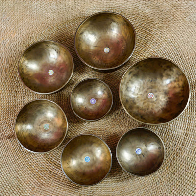 Seven handcrafted Himalayan singing bowls arranged together, featuring graduated sizes, chakra symbols, and warm antique bronze-gold finishes.