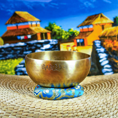 Front view of a handcrafted singing bowl with Om symbol, warm bronze finish, blue patterned cushion, and traditional wooden mallet.