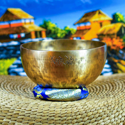 Front view of a handcrafted singing bowl with Om symbol, warm bronze finish, blue patterned cushion, and traditional wooden mallet.