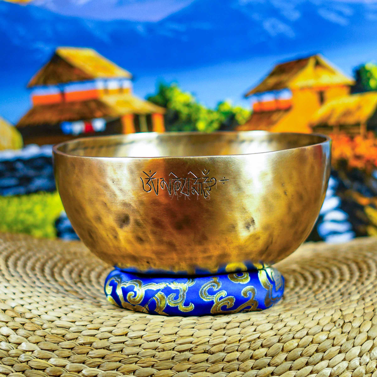 Front view of a handcrafted singing bowl with Om symbol, warm bronze finish, blue patterned cushion, and traditional wooden mallet.