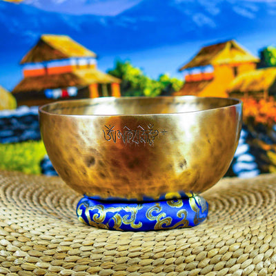 Front view of a handcrafted singing bowl with Om symbol, warm bronze finish, blue patterned cushion, and traditional wooden mallet.