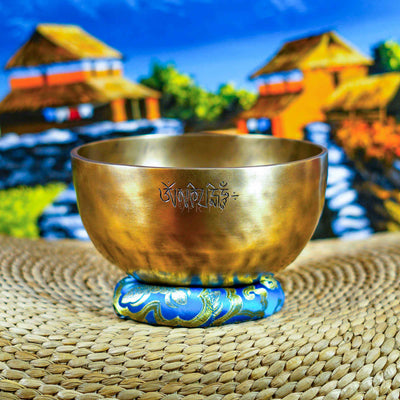 Front view of a handcrafted singing bowl with Om symbol, warm bronze finish, blue patterned cushion, and traditional wooden mallet.