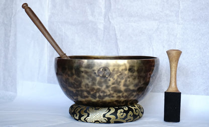 Hand-hammered brass singing bowl with a wooden striker and a red-padded mallet, displayed on a decorative cushion against a white background.