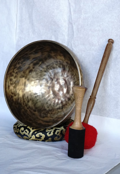 Hand-hammered brass singing bowl with a wooden striker and a red-padded mallet, displayed on a decorative cushion against a white background.