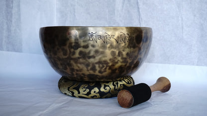 Hand-hammered brass singing bowl with a wooden striker and a red-padded mallet, displayed on a decorative cushion against a white background.