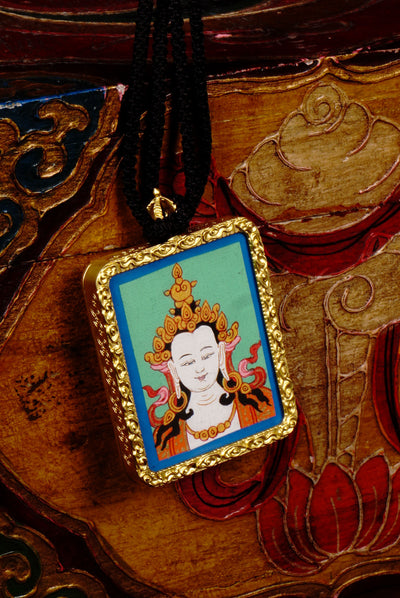 Square gold-framed Vajrasattva pendant with blue background, held in an open hand against a black backdrop.
