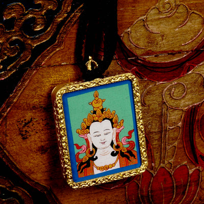 Square gold-framed Vajrasattva pendant with blue background, held in an open hand against a black backdrop.
