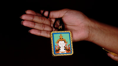 Square gold-framed Vajrasattva pendant with blue background, held in an open hand against a black backdrop.
