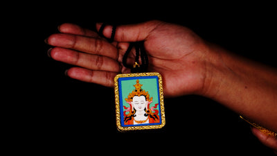 Square gold-framed Vajrasattva pendant with blue background, held in an open hand against a black backdrop.
