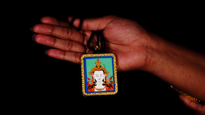 Square gold-framed Vajrasattva pendant with blue background, held in an open hand against a black backdrop.
