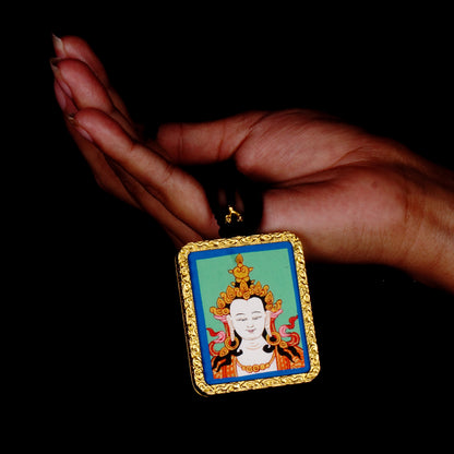 Square gold-framed Vajrasattva pendant with blue background, held in an open hand against a black backdrop.
