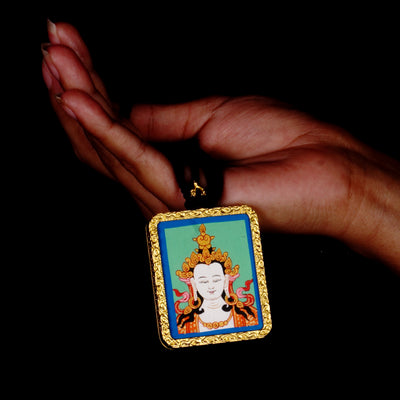 Square gold-framed Vajrasattva pendant with blue background, held in an open hand against a black backdrop.
