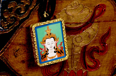 Square gold-framed Vajrasattva pendant with blue background, held in an open hand against a black backdrop.
