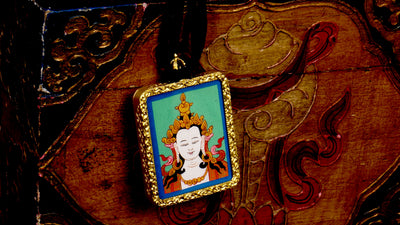 Square gold-framed Vajrasattva pendant with blue background, held in an open hand against a black backdrop.
