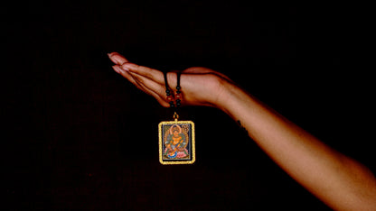 Hand-painted Zambala miniature Thangka pendant in gold-accented copper Ghau locket displayed hanging from black beaded cord.