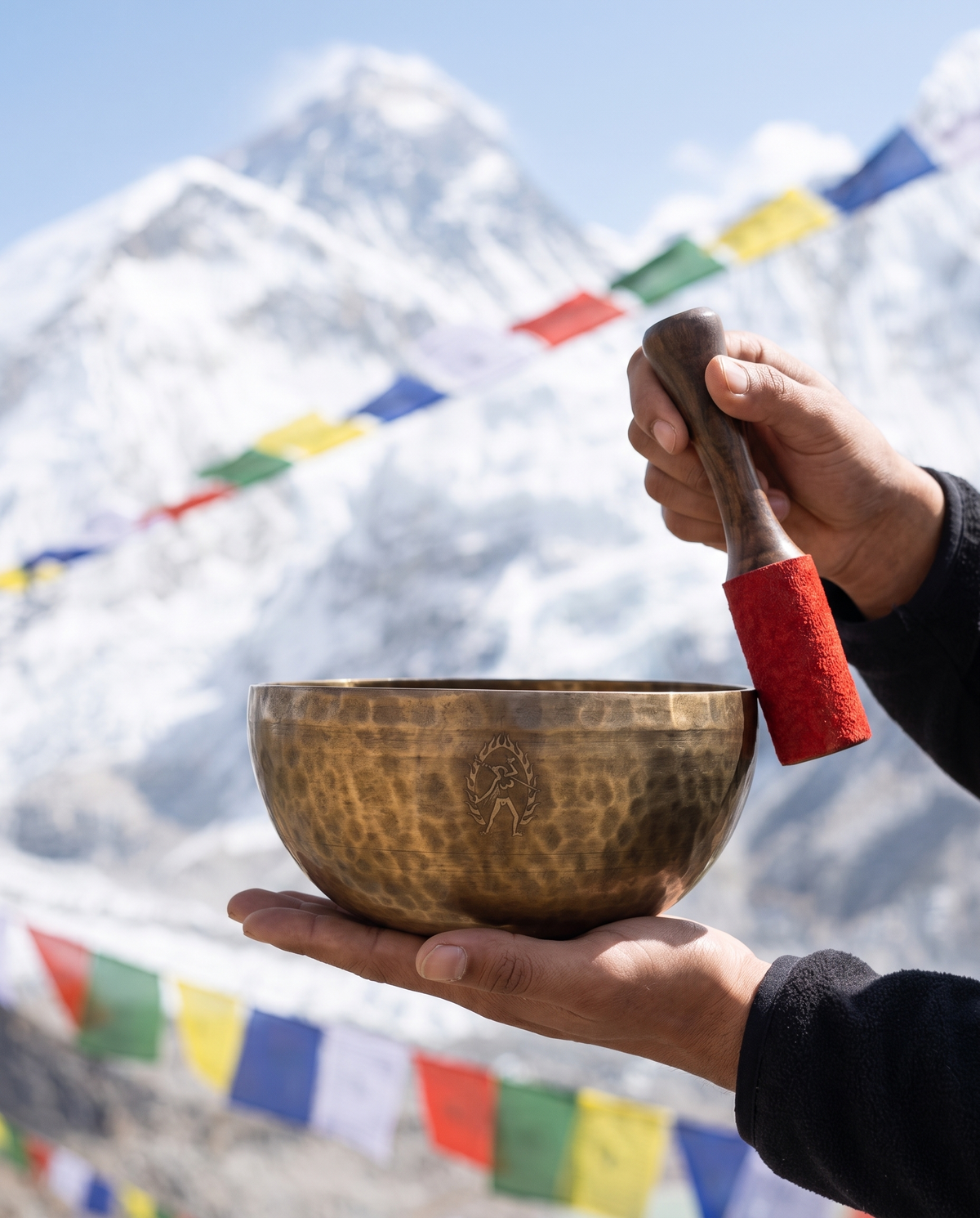 Handmade hammered Tibetan singing bowl and mallet held against snowy Himalayan peaks and colorful prayer flags