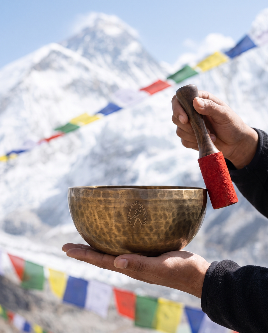 Handmade hammered Tibetan singing bowl and mallet held against snowy Himalayan peaks and colorful prayer flags