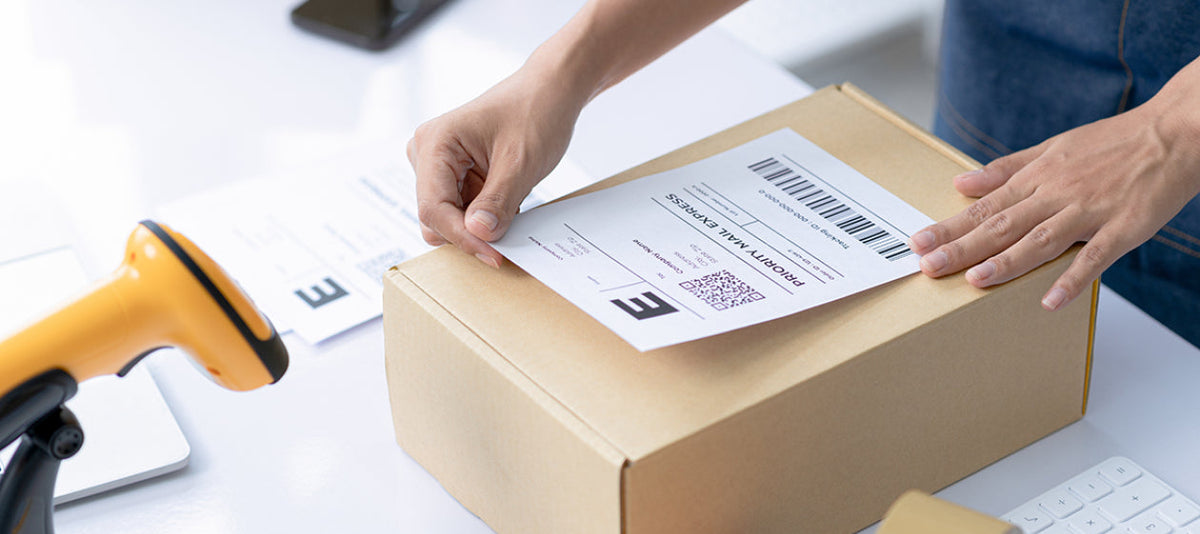 Hands pressing a courier label onto a sealed kraft shipping box, beside a barcode scanner and a roll of tape.
